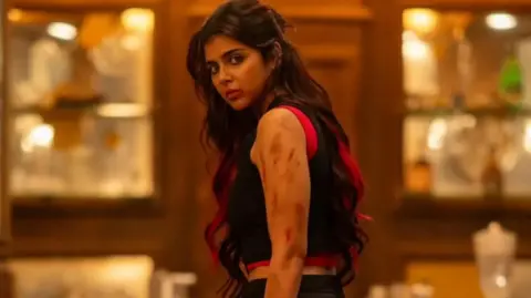 Wayfarer Films Actor Kalyani Priyadarshan seen in a red and black vest with bruises on her arm. She has red highlights in her hair.