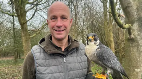 Lloyd stands in a woodland looking at the camera. He is of tall, bald with light stubble. He wears a brown fleece and grey gillet. A white and grey peregrine falcon sits on his fist.